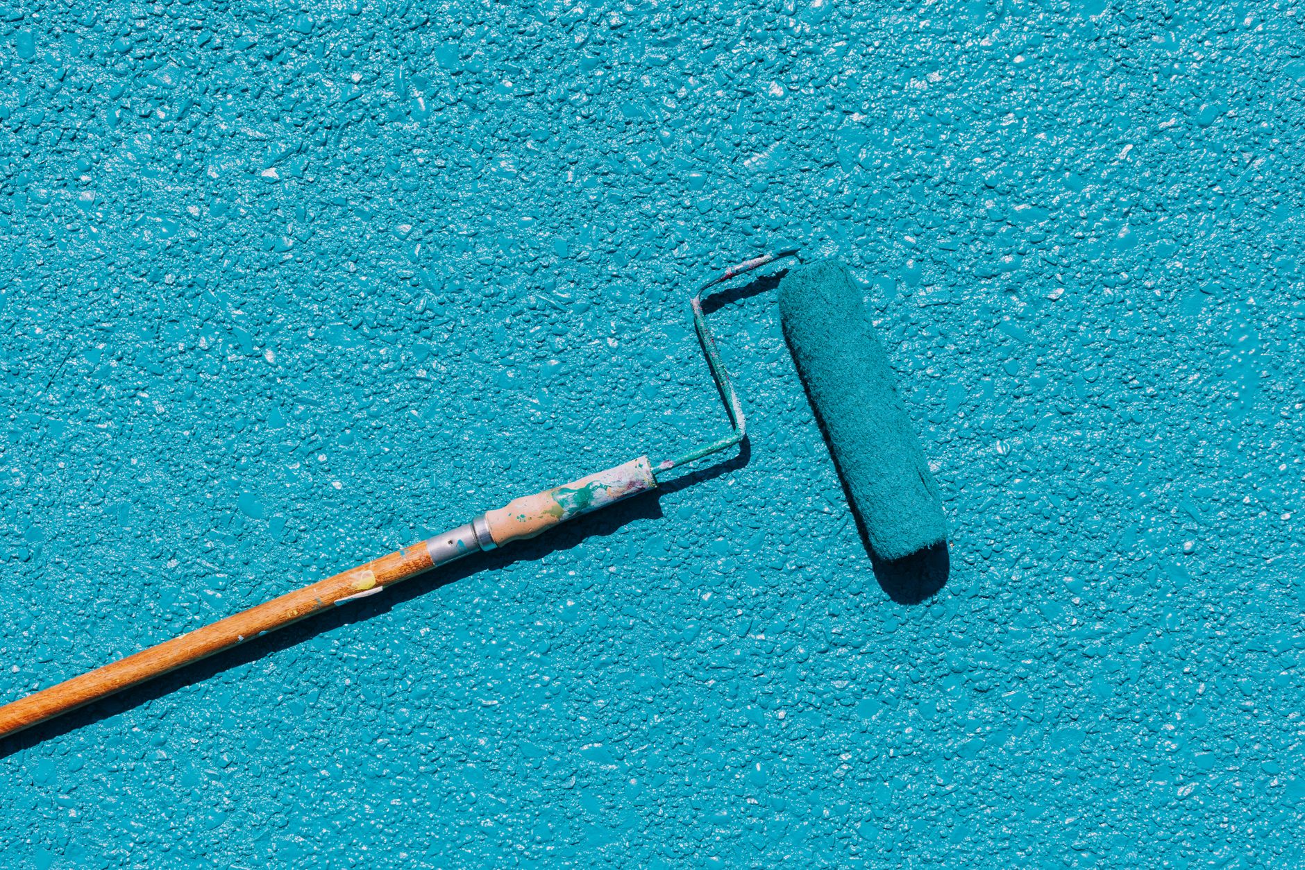 A close-up of a blue paint roller applying paint on a textured wall surface, captured in bright sunlight in Carlsbad, CA